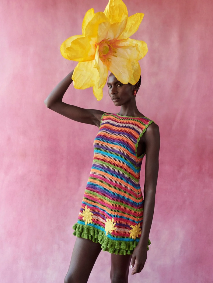 Model wearing a colorful striped dress with a large yellow flower headpiece against a pink background