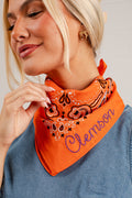 A close-up lifestyle shot of a woman wearing a bright orange bandana with a paisley pattern and the name 'Clemson' embroidered in purple script. She is wearing a blue denim top against a light gray background.