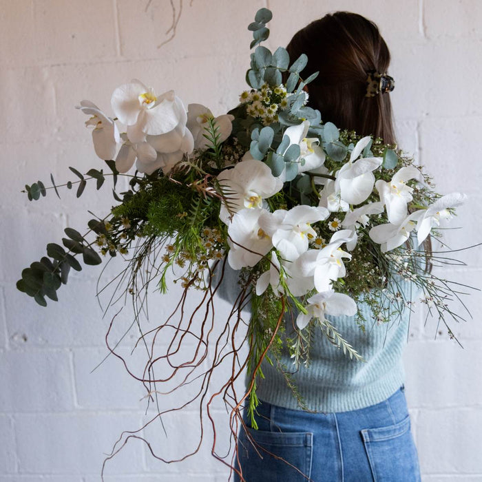 Person holding a large bouquet of white orchids and greenery against a white wall.