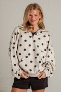 A woman wearing a cream-colored half-zip hoodie with black polka dots, paired with black shorts, standing against a light gray brick wall.