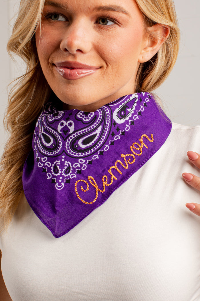 A close-up of a woman wearing a purple bandana bib with a paisley pattern and the name 'Clemson' embroidered in orange on the front.
