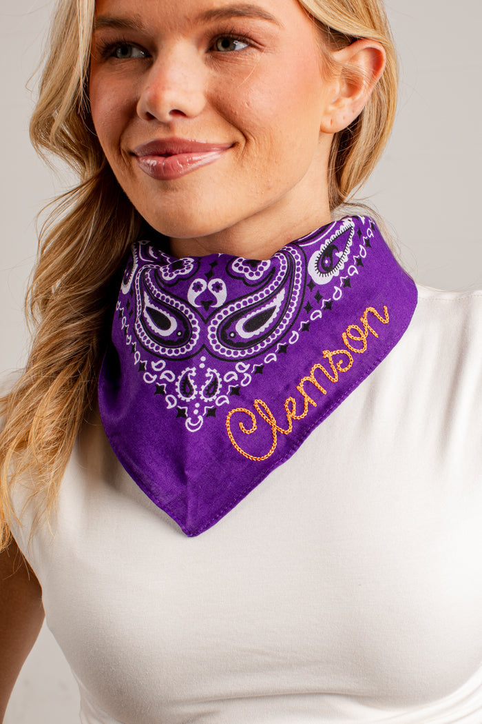 A close-up of a woman wearing a purple bandana-style neck gaiter with white paisley embroidery and the name 'Clemson' stitched in orange script.
