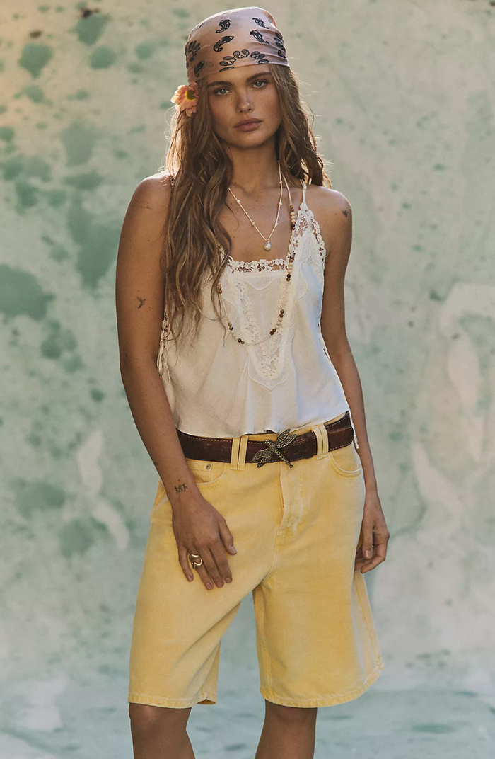 Woman wearing a ivory top and yellow shorts with a floral headband against a textured wall.