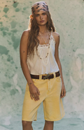 Woman wearing a ivory top and yellow shorts with a floral headband against a textured wall.