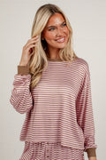 A smiling blonde woman wearing a long-sleeve dusty rose and white striped top with brown ribbed cuffs, standing against a light off-white brick wall.