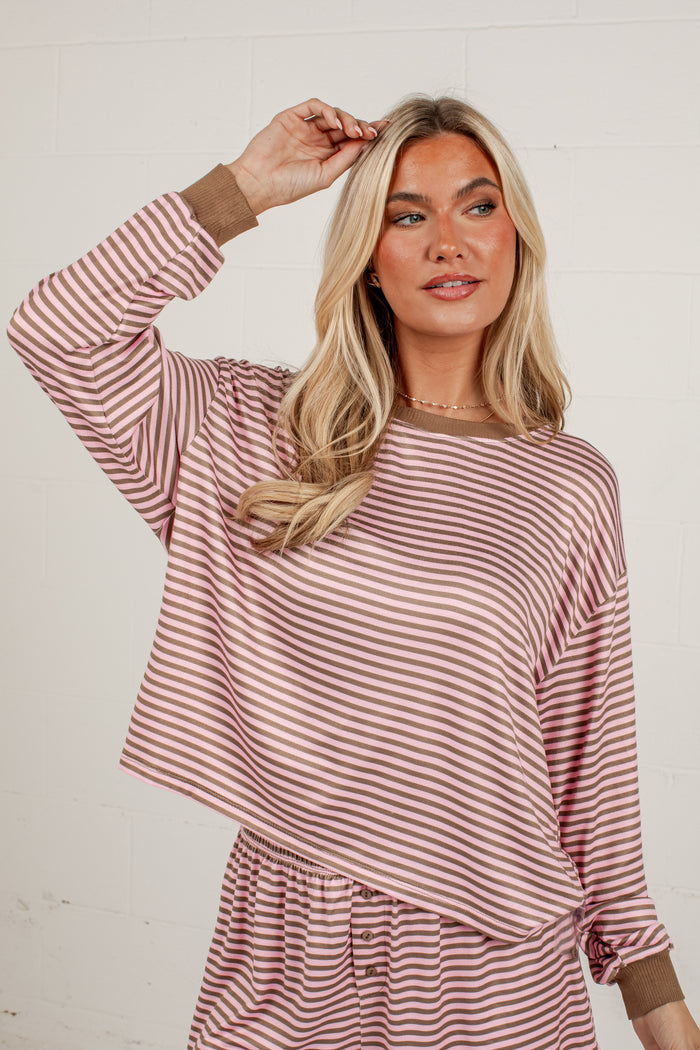 A woman modeling a long-sleeve striped top with a dusty rose and white pattern, featuring solid brown ribbed cuffs and collar, set against a white brick wall background.