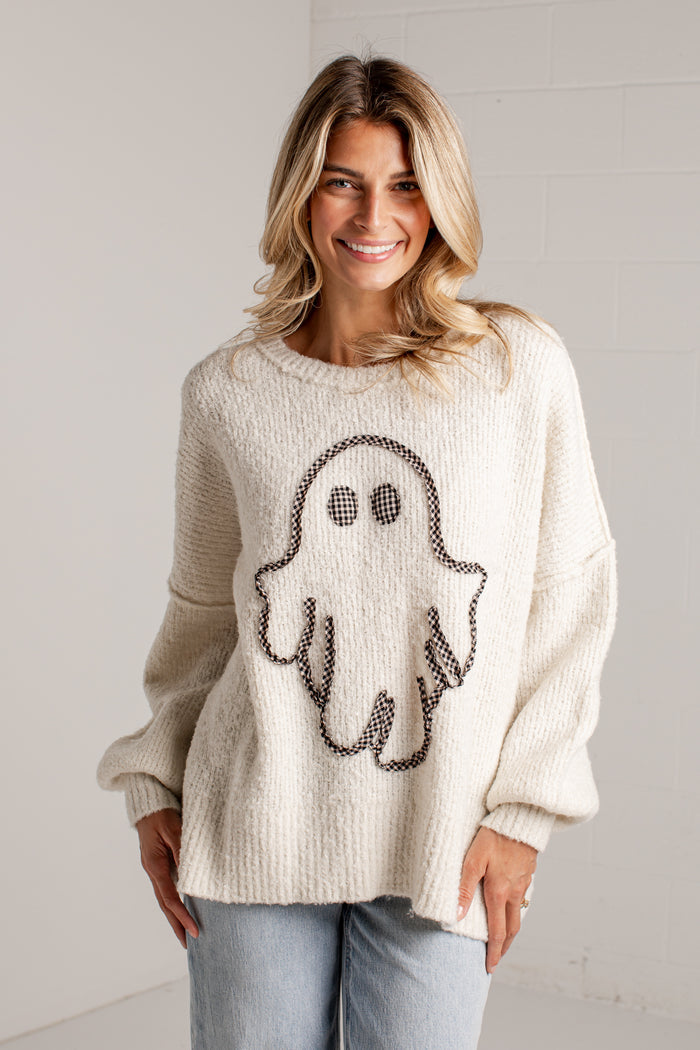 A woman wearing a cream-colored knit sweater with a black ghost graphic on the front, paired with light blue jeans, standing against a white brick wall.