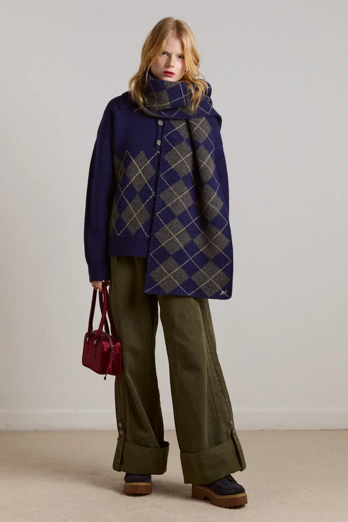 Woman wearing a blue coat with a plaid pattern and Wylie Turn Up Jeans in khaki holding a red bag against a plain background